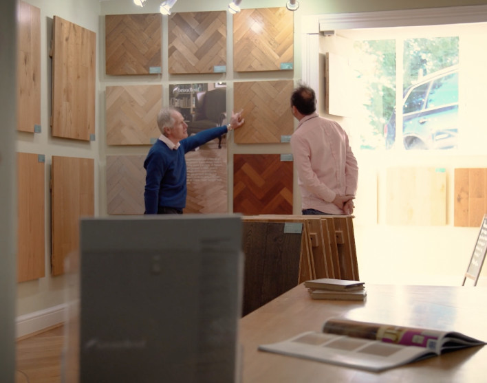 A broadleaf flooring expert guides a customer around a Broadleaf showroom, indicating to samples mounted on the walls.